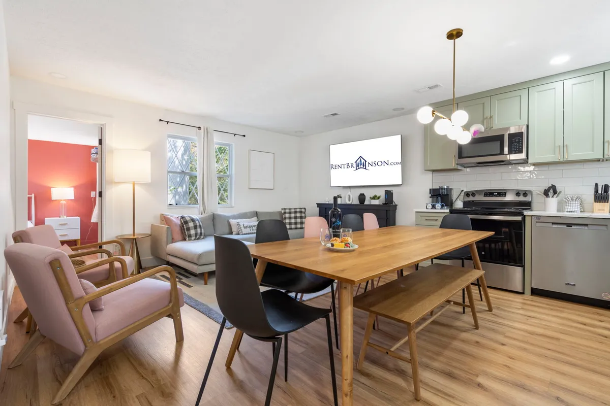 The image showcases the inviting heart of the home—an open-concept living and dining area featuring a spacious dining table perfect for family meals, a cozy sofa for relaxing evenings, a television for entertainment, and large windows that fill the space with natural light. The adjoining kitchen area hints at easy meal prep and gathering with loved ones.