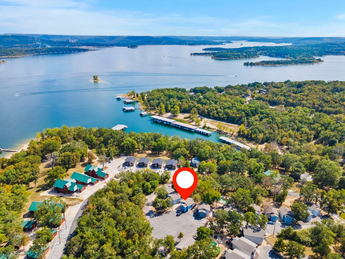This exterior scene captures a tranquil lakeside setting, with cabins nestled among trees, a dock extending towards the water, and distant views of people boating and enjoying nature. The atmosphere evokes lakeside leisure and outdoor fun.