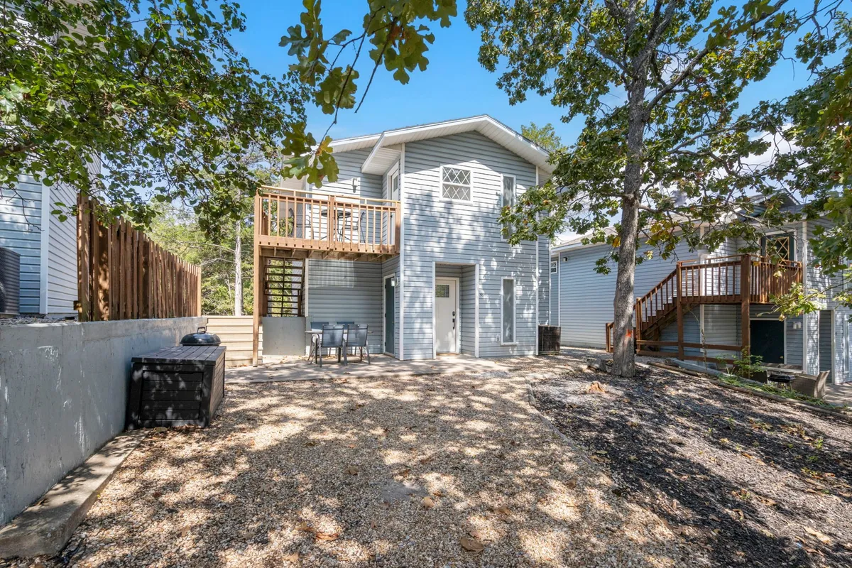 An inviting deck set amidst landscaped greenery and trees, with a gravel path leading away and comfortable outdoor seating visible. The scene suggests relaxation, outdoor dining, and a seamless connection to the natural environment.