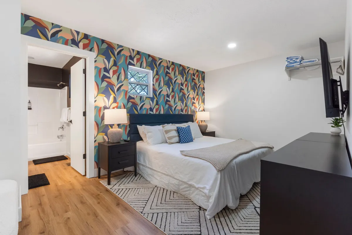 A serene bedroom featuring a plush bed with a floral accent wall, nightstands on either side, and a flat-screen television. The decor is soft and inviting, ideal for winding down after a busy day. Natural light gently filters into the room, complementing the soothing atmosphere.