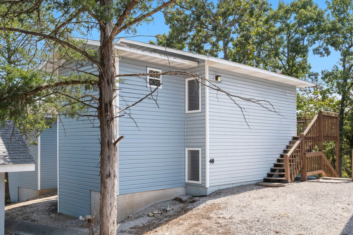 A welcoming exterior view that highlights a staircase leading to the deck, with tree branches overhead and the home’s exterior siding in view. The space hints at outdoor relaxation, barbecue potential, and access to the natural environment.