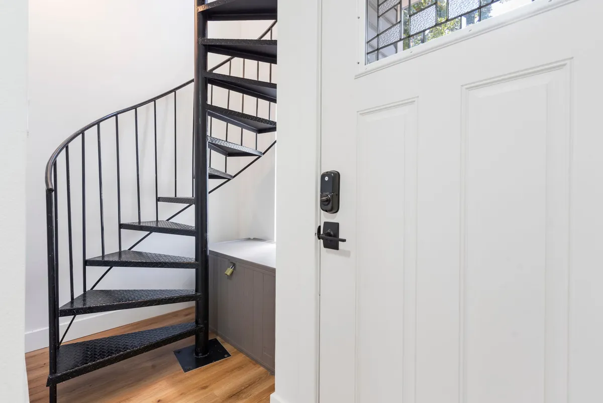 This image features a spiral staircase illuminated by natural light from a nearby window, with an entryway door in view. The staircase's whimsical design draws the eye and creates an inviting, eclectic first impression.