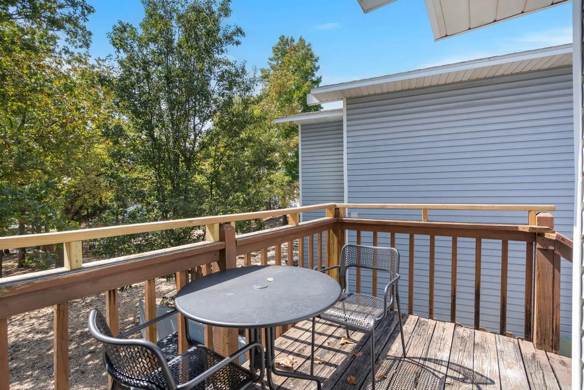 An inviting outdoor dining area featuring a table and chairs set on a deck, bordered by a railing and surrounded by leafy trees. The space suggests a setting for shared meals, morning coffee, or family gatherings in the fresh air.
