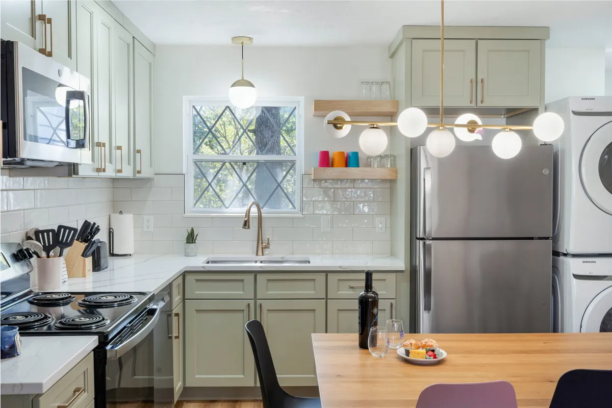 The photo centers on a spacious, modern kitchen with a prominent island topped by a stylish light fixture. Colorful cups on a shelf add a playful touch. The setting is ready for cooking, entertaining, or sharing meals, reinforcing the kitchen as a hub for culinary activity and shared experiences.