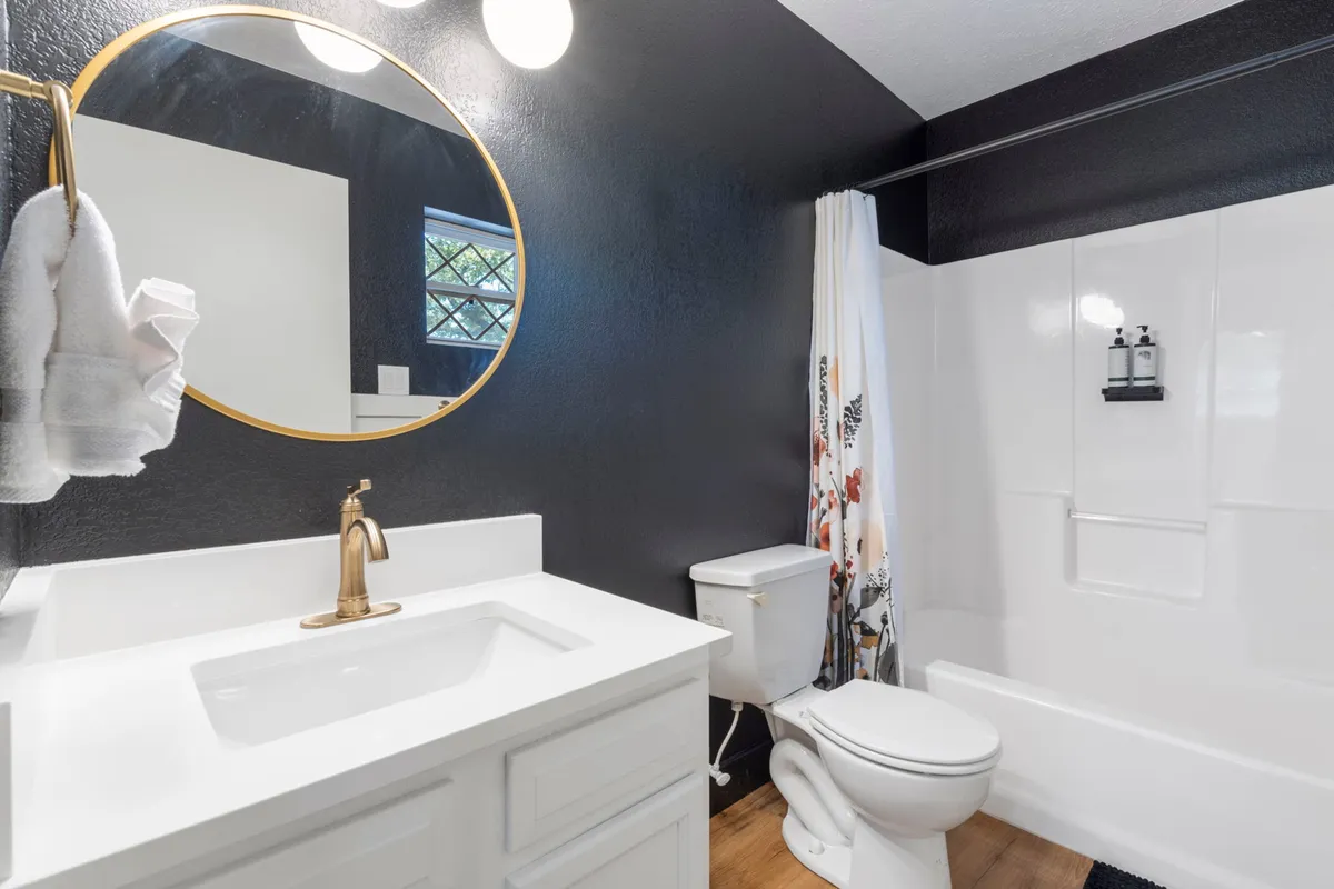 A bright, contemporary bathroom featuring a round mirror, gold faucet, floral shower curtain, and a white vanity. The atmosphere is fresh and clean, ideal for morning routines or relaxing evening baths.