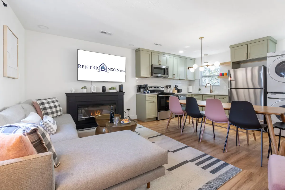 This image captures an open-concept living area featuring plush sofas gathered around a cozy fireplace, with a clear view into the kitchen and dining space. The streaming TV is visible, and the area is well-lit, exuding a modern yet inviting atmosphere. People can be seen socializing and relaxing, highlighting the space's versatility for gatherings and downtime.