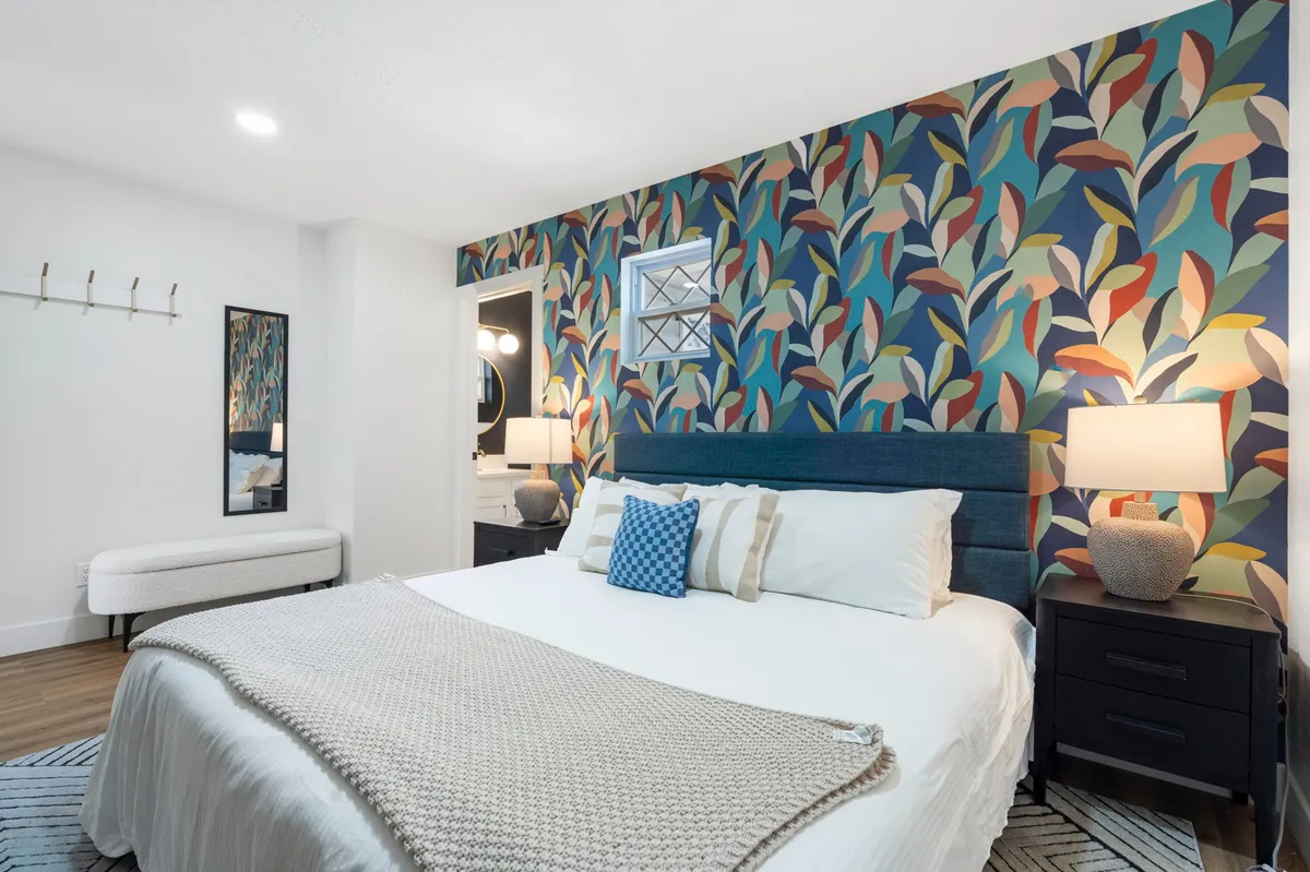 A vibrant bedroom interior with colorful, leaf-patterned wallpaper behind a bed adorned with decorative pillows and flanked by nightstands with lamps. The atmosphere is both lively and cozy, emphasizing eclectic and whimsical design choices.